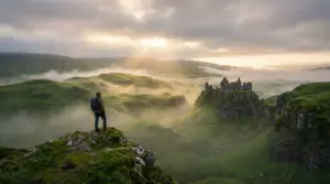 A lone traveler stands on a rocky hillside overlooking the misty Scottish Highlands